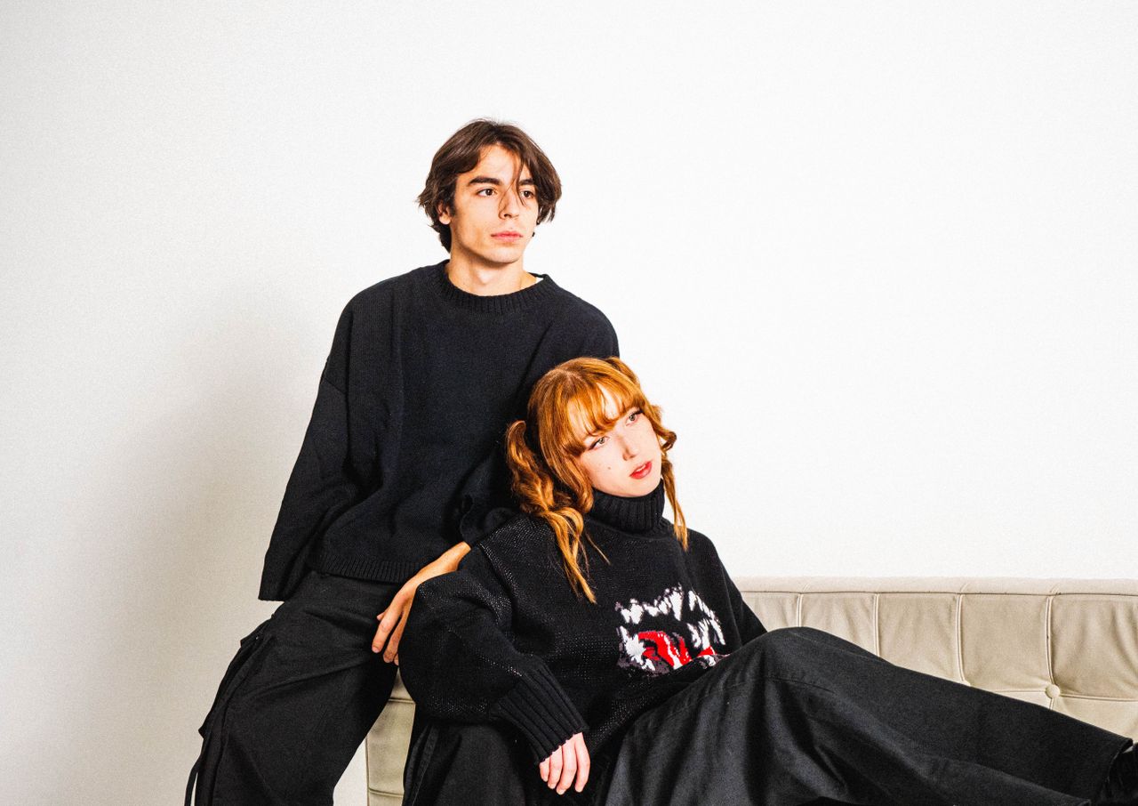 Home A fashionable couple dressed in black sits and poses thoughtfully on a couch.