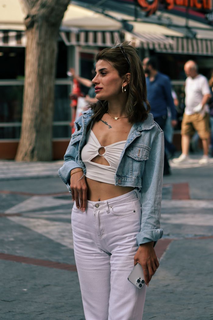 Services Stylish woman wears a jean jacket and holds a phone while standing outdoors on a city sidewalk.